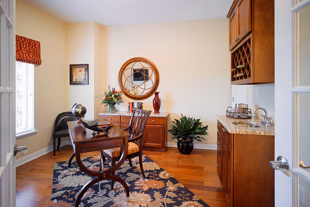 Bright furnished interior room with a wooden desk and chair, decorative rug, sideboard with mirror, plants, and a small wet bar/kitchenette.