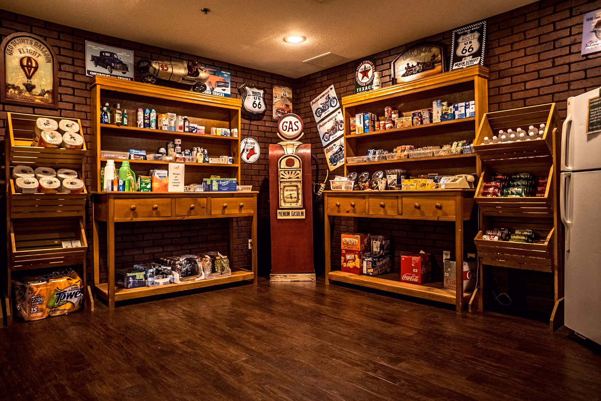 Interior of a small convenience store area with wooden shelves stocked with various grocery items, snacks, canned goods, and beverages. The walls are decorated with vintage Route 66 and Texaco signs, and there is a retro-style gas pump in the center. The floor is wooden, and a white refrigerator is visible on the right side.