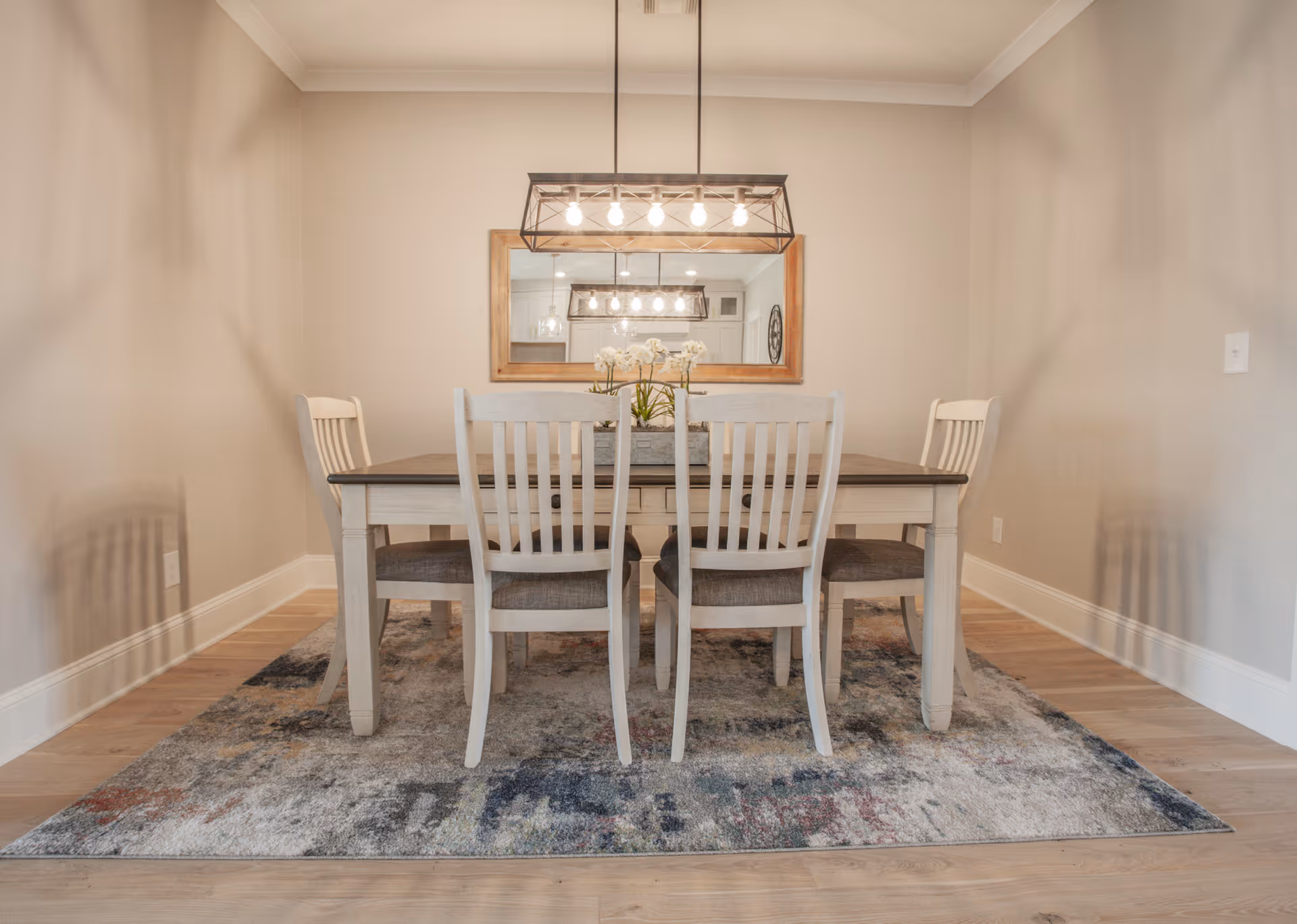 A dining room with a rectangular wooden table and six matching chairs with cushioned seats. Above the table hangs a rectangular light fixture with six exposed bulbs. A large mirror with a wooden frame is mounted on the wall behind the table, reflecting the light fixture. The floor is wooden with a patterned area rug under the table and chairs. The walls are painted a light beige color.