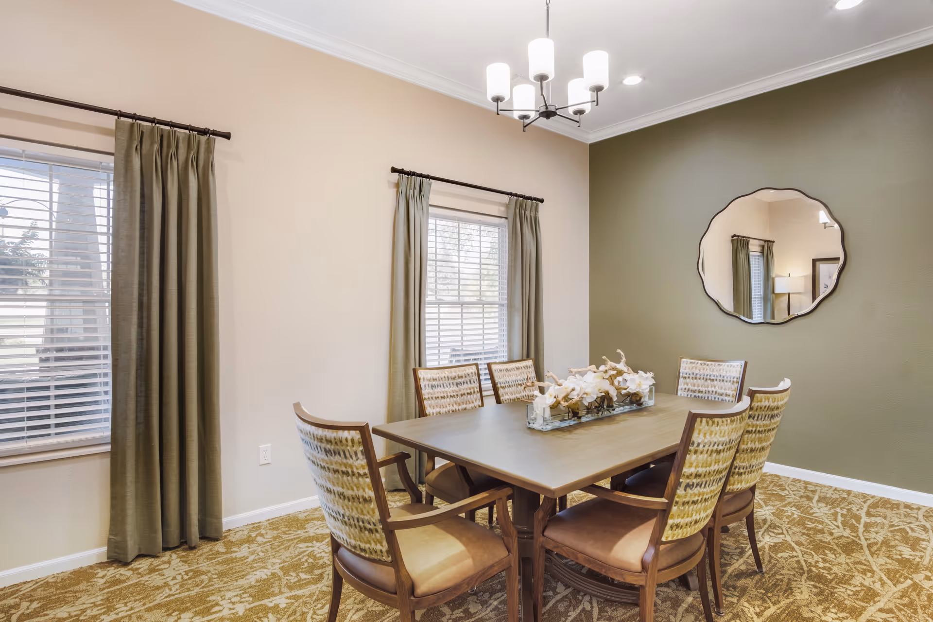 A dining room with a rectangular wooden table surrounded by six upholstered chairs, a chandelier overhead, green curtains on windows and a round mirror on a green accent wall.