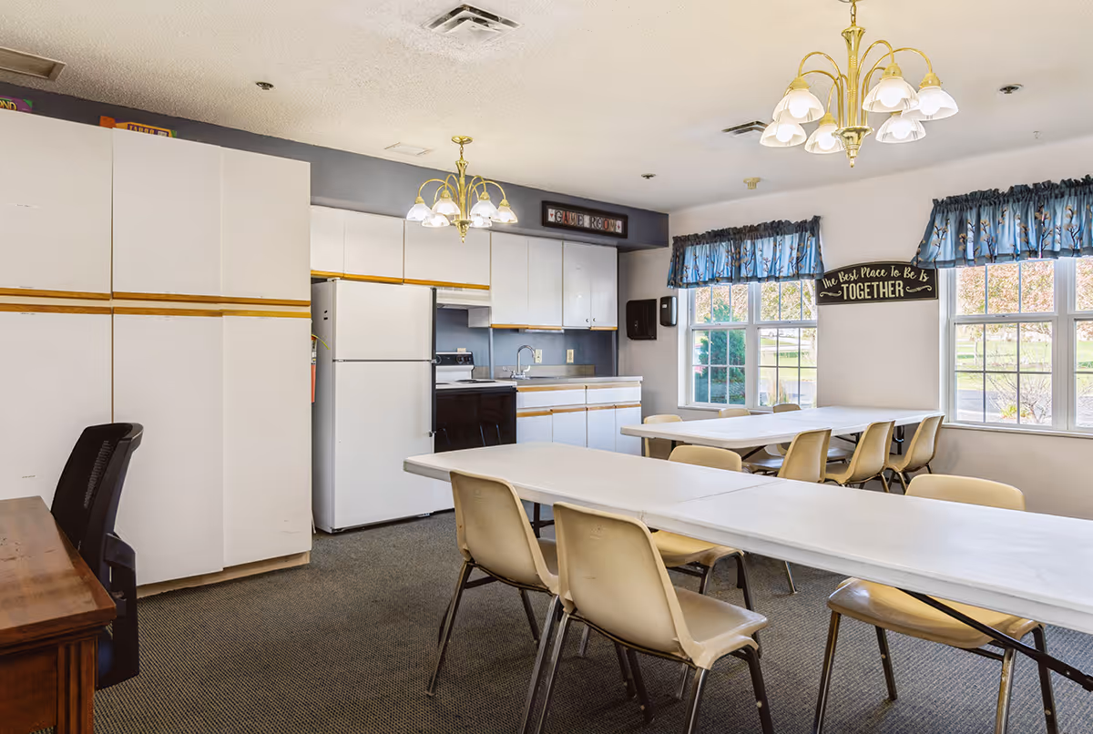 A communal dining/activity room with folding tables and chairs, a kitchenette with white cabinets and appliances, and windows with blue valances.