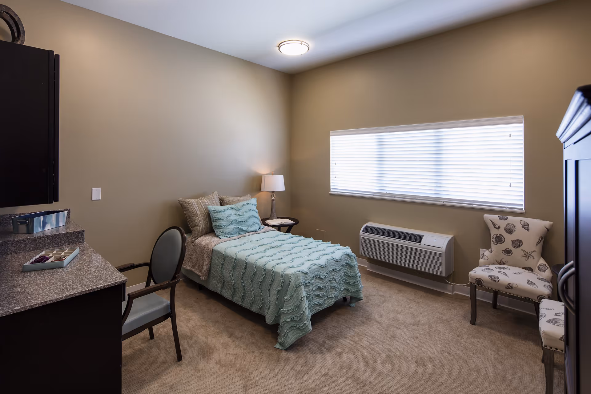 Small furnished bedroom with a single bed, bedside table and lamp, two chairs, and a window with blinds.