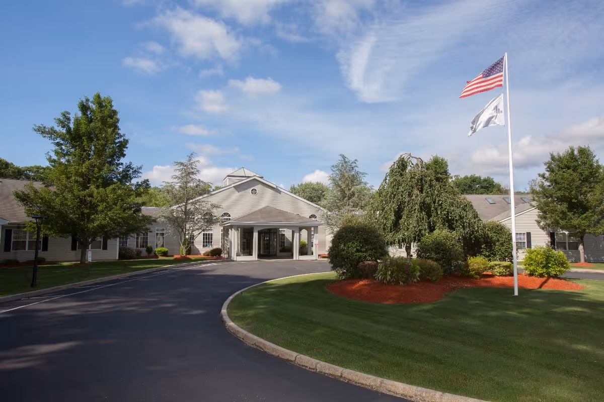 Exterior view of The Village at East Farms facility with a curved driveway leading to the main entrance. The building is surrounded by well-maintained landscaping including trees, bushes, and a lawn. Two flags, one American and one white, are flying on a flagpole near the entrance under a partly cloudy blue sky.