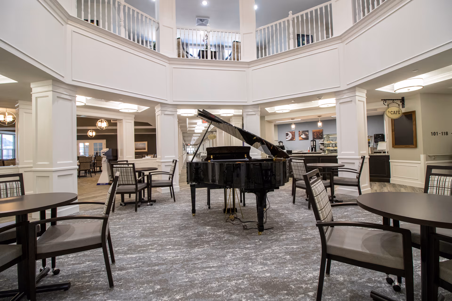 Interior view of a senior living facility common area featuring a black grand piano in the center, surrounded by round tables and chairs. The space has white pillars and railings with a second-floor balcony overlooking the area. In the background, there is a café counter with coffee machines and a sign that reads 'CAFÉ'.