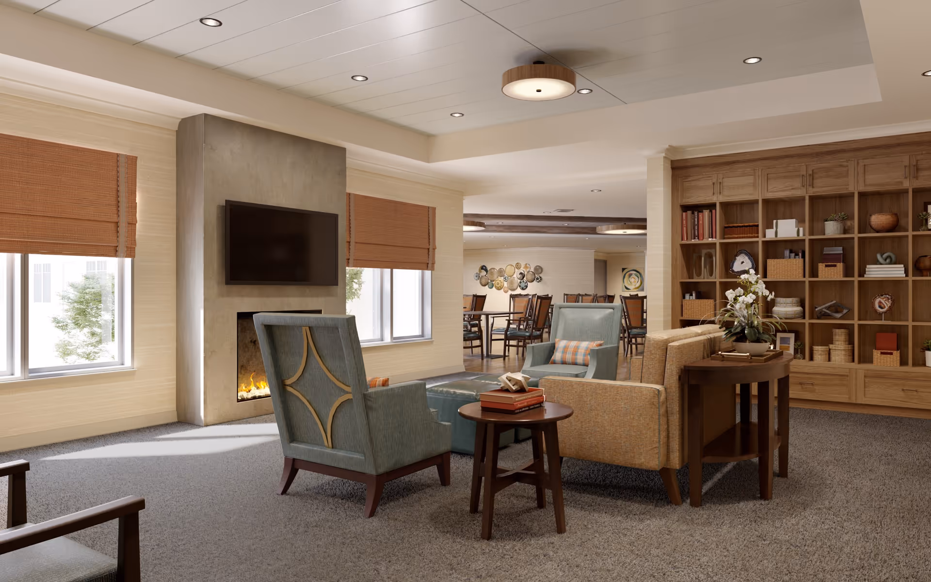 A cozy living room area in Sunrise of Redmond featuring a modern fireplace with a mounted flat-screen TV above it, two blue armchairs with patterned cushions, a beige sofa, a small round wooden side table with books, and a large wooden bookshelf filled with decorative items and books. The room has large windows with brown blinds, carpeted flooring, and recessed ceiling lights. In the background, there is a dining area with tables and chairs.