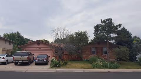 Single-story brick house front with a driveway holding parked cars, a lawn and trees under a cloudy sky.