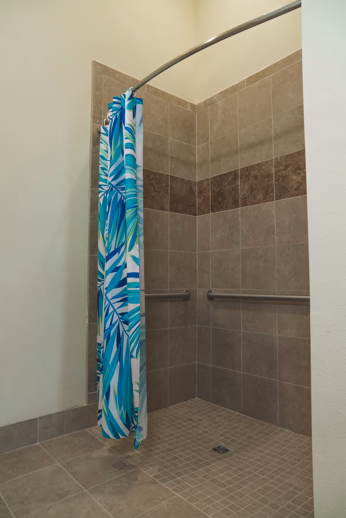 A tiled walk-in shower with beige and brown tiles, featuring two horizontal grab bars on the walls and a curved shower curtain rod with a blue and green patterned shower curtain partially drawn.