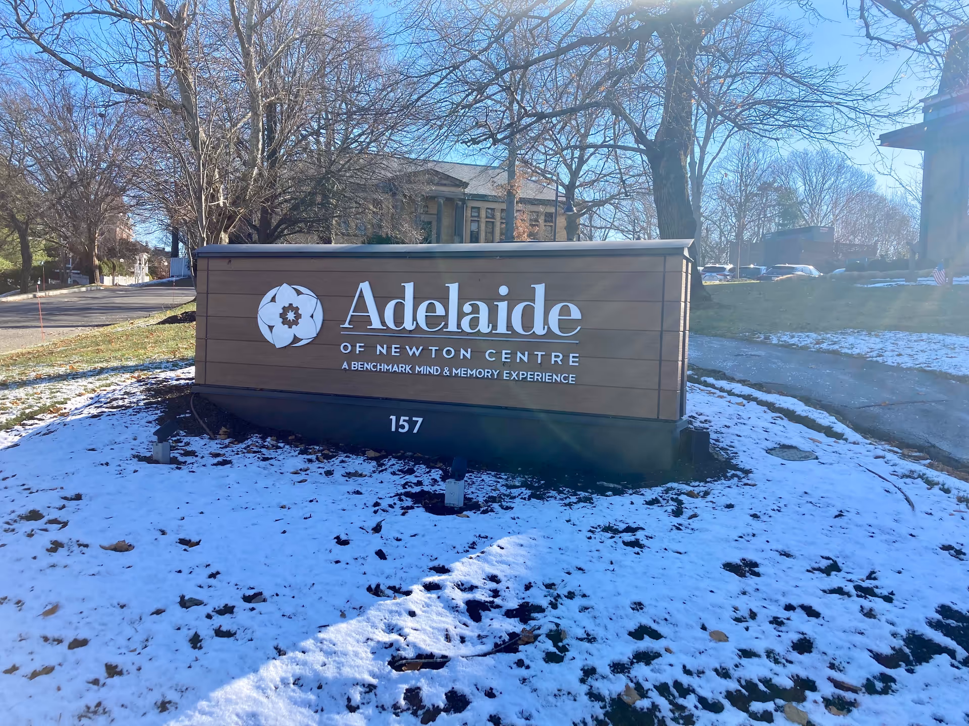 Outdoor view of a large wooden sign for Adelaide of Newton Centre, a Benchmark Mind & Memory Experience, with the address number 157. The ground around the sign is covered with patches of snow, and leafless trees and a building are visible in the background under a clear blue sky.