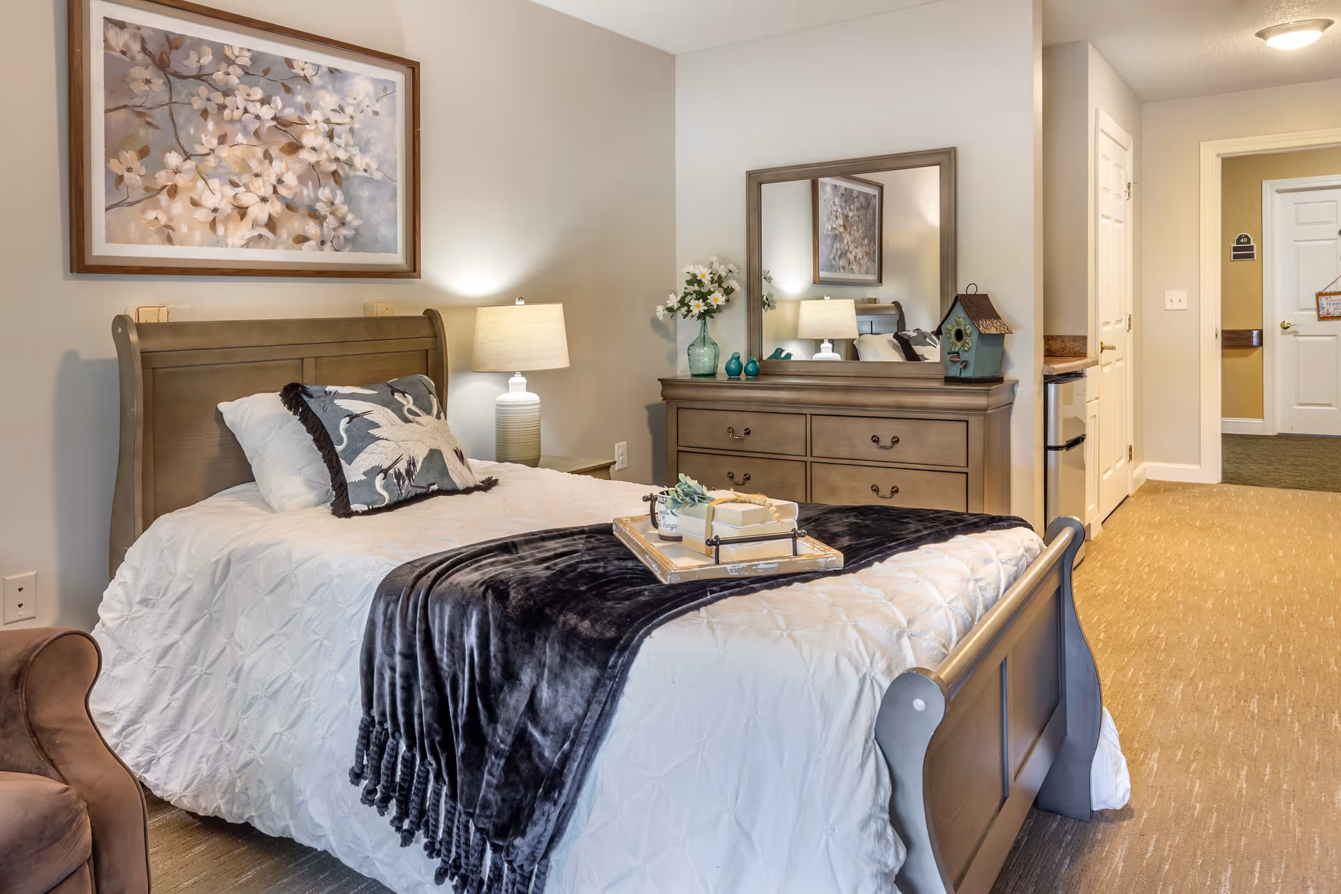 A cozy bedroom with a wooden bed frame, white bedding, and a dark gray throw blanket. There is a decorative pillow with a bird design on the bed. A wooden dresser with a large mirror is against the wall, adorned with a vase of flowers and small decorative items. A table lamp is on a nightstand beside the bed. The room has beige walls and carpet, with a framed floral painting hanging above the bed. A hallway with a white door is visible in the background.