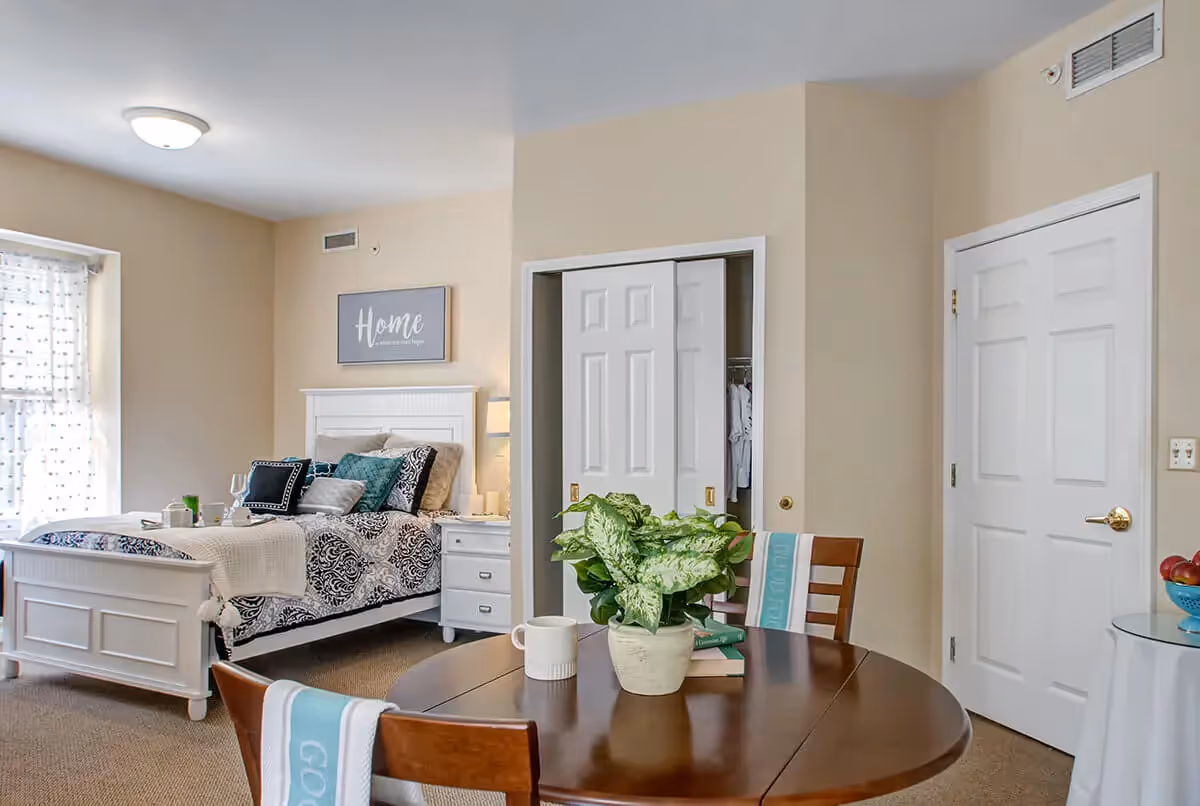 A cozy bedroom with a white bed frame and patterned bedding, several pillows, a nightstand with a lamp, and a wall sign that reads 'Home is where our story begins.' In the foreground, there is a round wooden table with a potted plant, a coffee mug, and two chairs with light blue towels draped over them. The room has beige walls, a window with sheer curtains, a closet with sliding doors, and a closed white door.