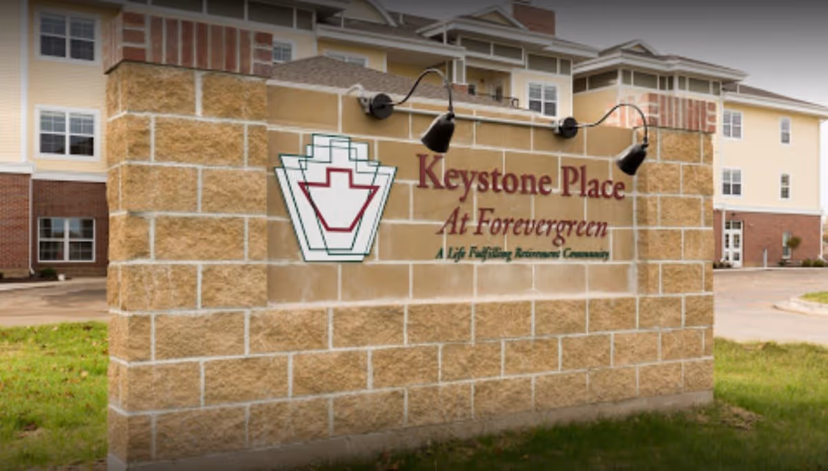 A large stone sign outside a building displaying the name 'Keystone Place At Forevergreen' with the tagline 'A Life Fulfilling Retirement Community'. The building in the background is a multi-story structure with multiple windows and a brick and beige exterior.