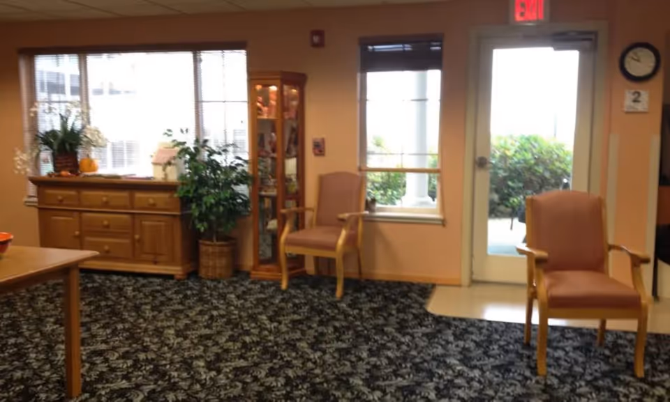 A cozy senior living common room with upholstered chairs, a wooden cabinet and display case, potted plants, patterned carpet, and a door leading outside.