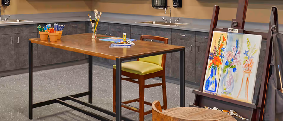 Art studio area with a wooden table holding paintbrushes, colored pencils, and art supplies. There is a chair with a yellow cushion at the table. An easel displays a colorful painting of flowers in vases. The background shows gray cabinets with sinks and faucets.