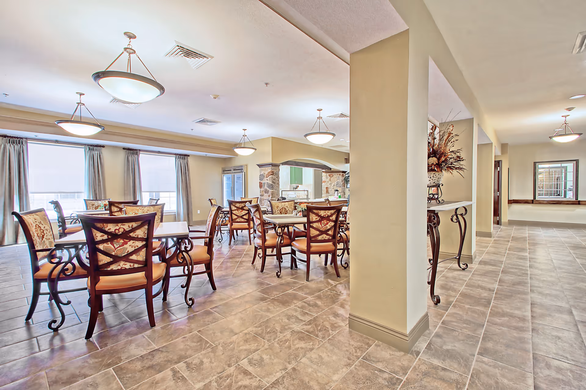 Bright communal dining area with multiple tables and upholstered chairs, pendant lights, tiled floor, and a serving counter.