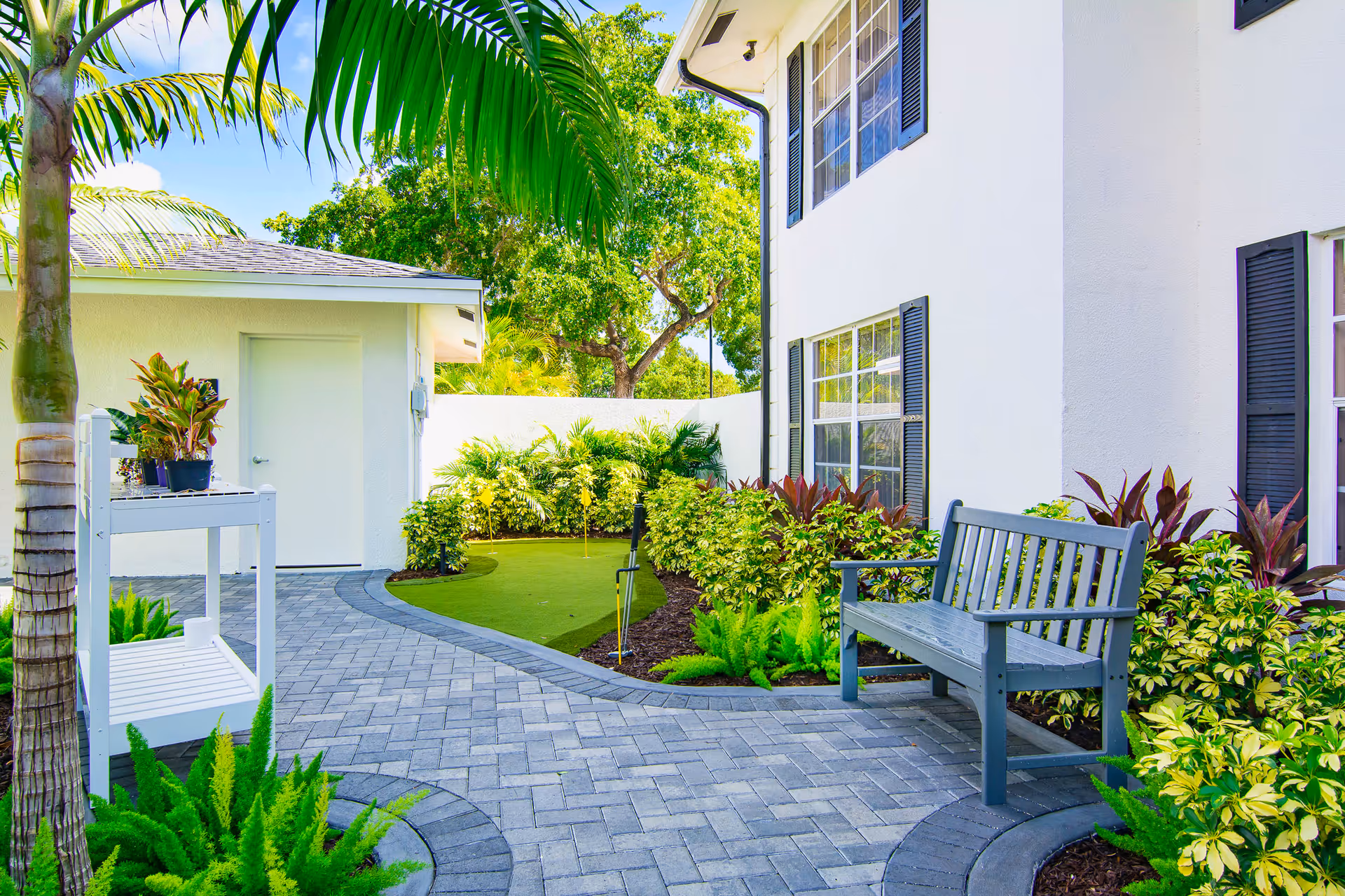 A bright outdoor patio area with a paved walkway, a gray wooden bench, lush green plants, and a small putting green surrounded by shrubs. The side of a white building with black window shutters is visible, along with a small white structure with a closed door. Palm trees and other greenery are in the background under a clear blue sky.