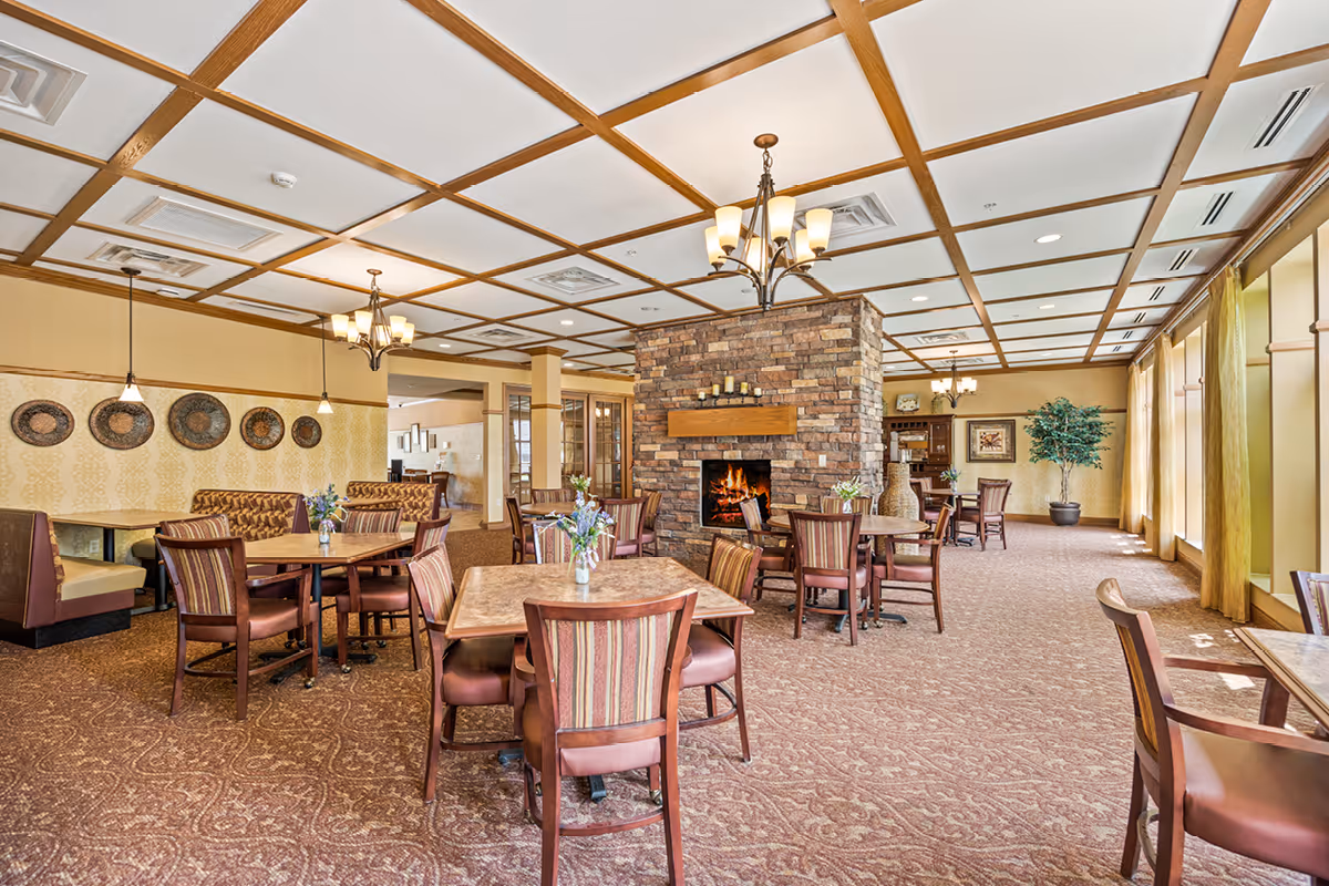 A spacious dining room with multiple wooden tables and chairs arranged around a central stone fireplace. The room features a coffered ceiling with chandeliers, large windows with yellow curtains allowing natural light, and decorative wall art. The carpet has a patterned design, and there are some plants and vases with flowers placed around the room.