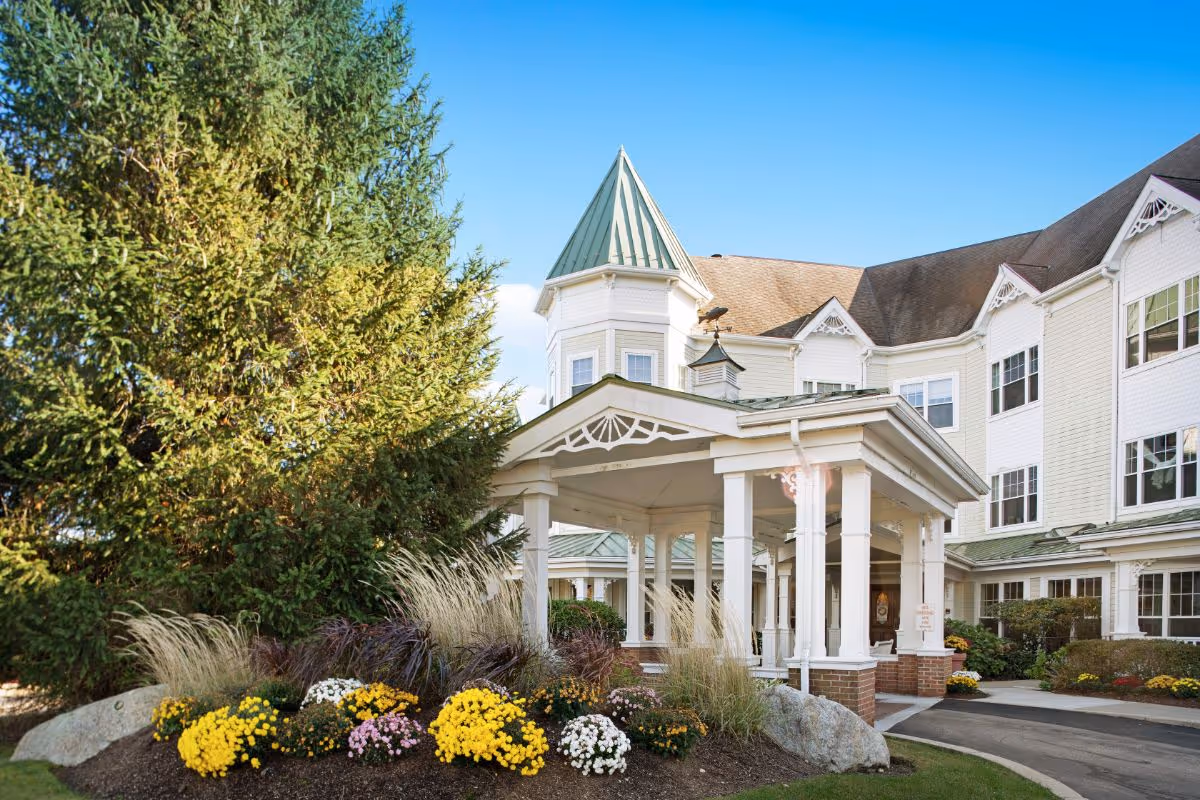 Front entrance and covered portico of a senior living building with a turret, landscaped flower beds and a large evergreen tree.