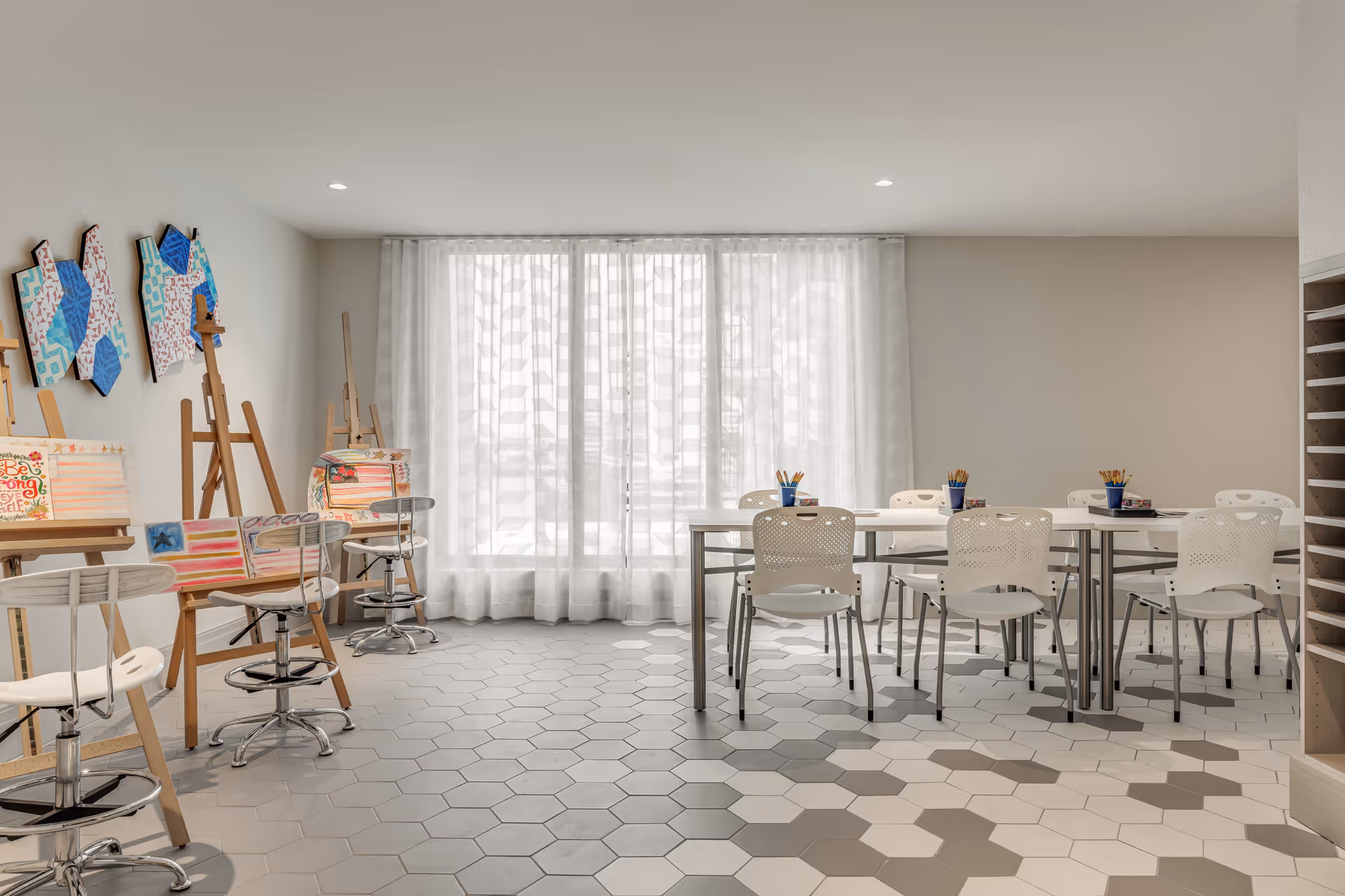 Bright activity room with easels, art supplies, tables and chairs in front of a large curtained window.