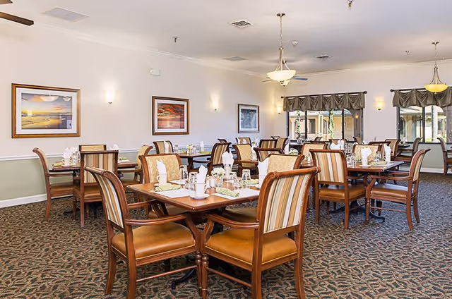 A well-lit dining room with multiple wooden tables and chairs arranged neatly. Each table is set with white napkins, glasses, and silverware. The room has patterned carpet flooring, framed artwork on the walls, and large windows with valance curtains allowing natural light to enter.