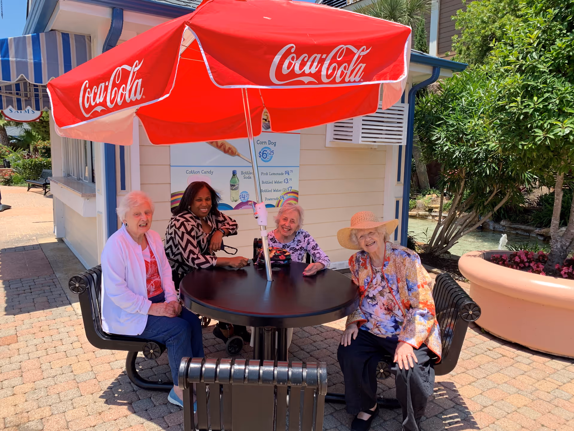 Four elderly women sitting around a black outdoor table under a red Coca-Cola umbrella. They are smiling and enjoying a sunny day outside near a small building with a menu sign. There are plants and a large planter visible in the background.