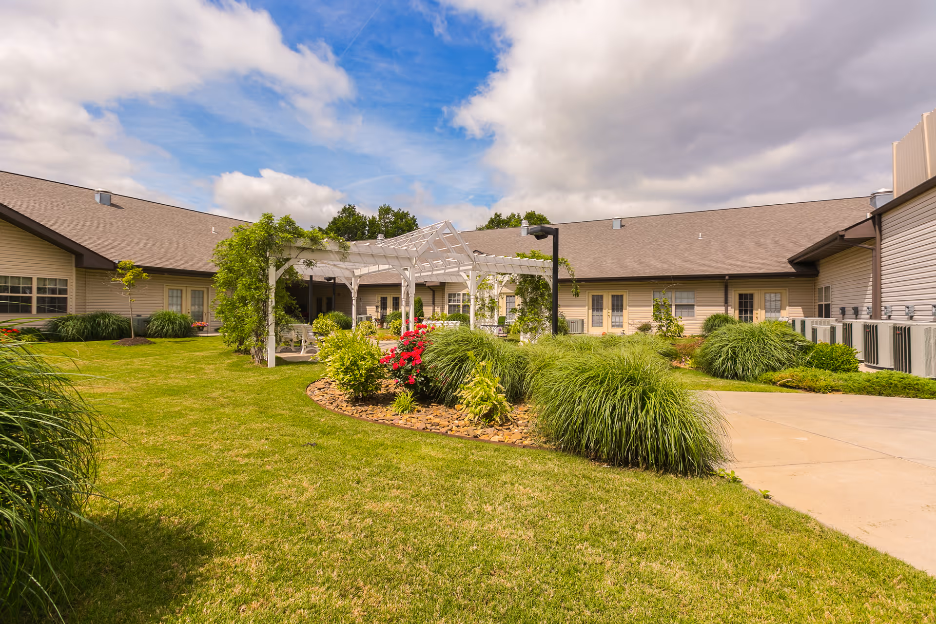 Sunlit courtyard with a manicured lawn, flower beds, a white pergola, and a surrounding single-story senior living building.