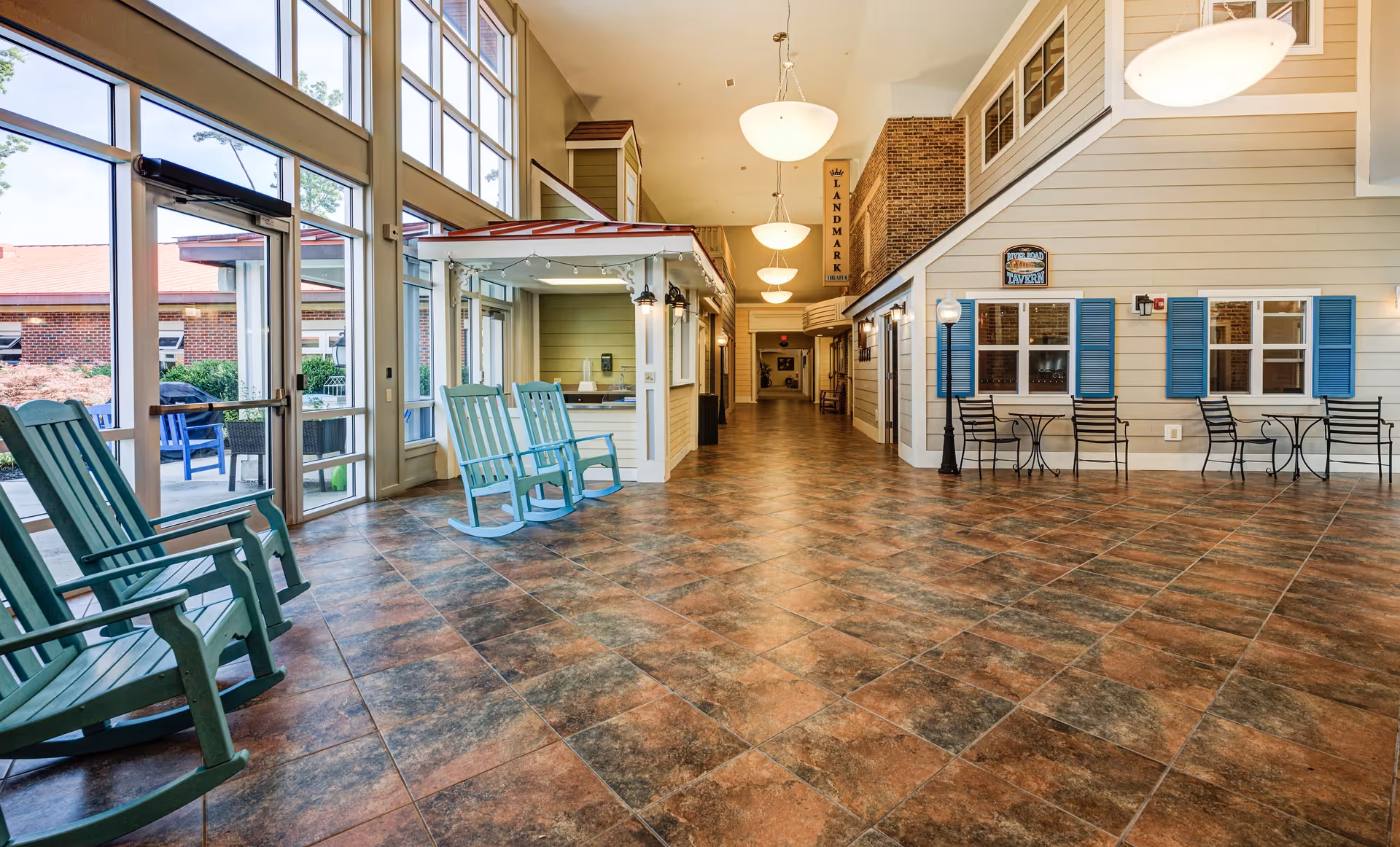 Spacious indoor common area with large windows letting in natural light, featuring green rocking chairs on the left, a small kiosk or reception area in the center, and a wall designed like a house facade with blue shutters and a sign reading 'Tavern' on the right. The floor is tiled with a brown and gray pattern, and there are hanging light fixtures along the ceiling.