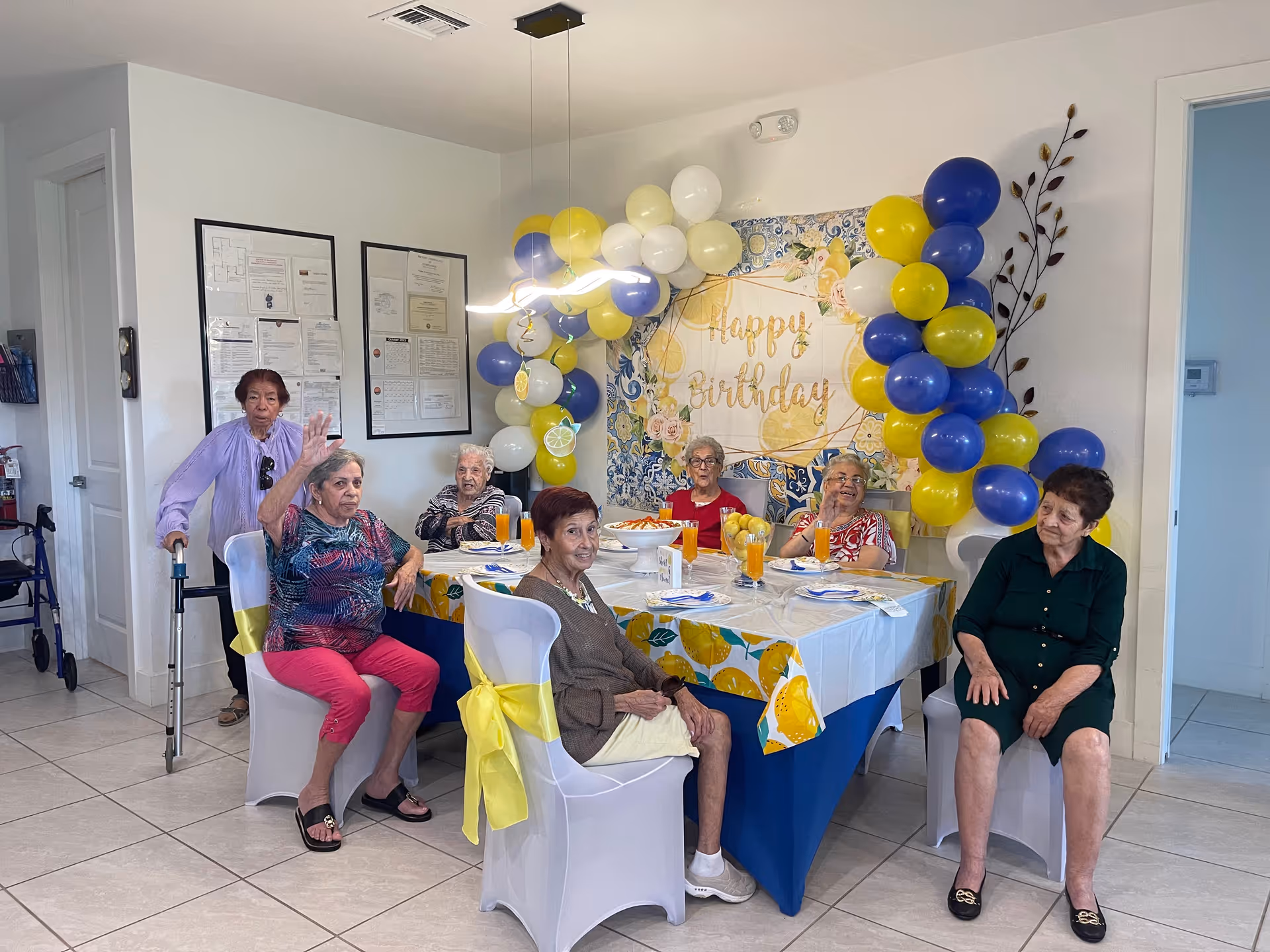 A group of elderly women gathered around a table decorated with a lemon-themed tablecloth and yellow, white, and blue balloons. A 'Happy Birthday' banner is displayed on the wall behind them. The women are seated on white chairs with yellow bows, and there are glasses of orange drink on the table. The room has white walls and tiled floors.