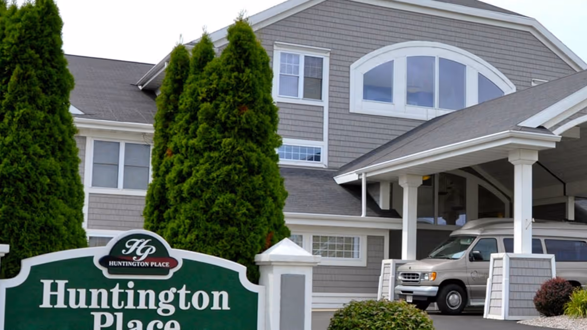Exterior view of Huntington Place senior living facility showing a gray building with white trim, large windows, a covered entrance, and a parked van. There are tall green trees and a green sign with white text reading 'Huntington Place' in front.