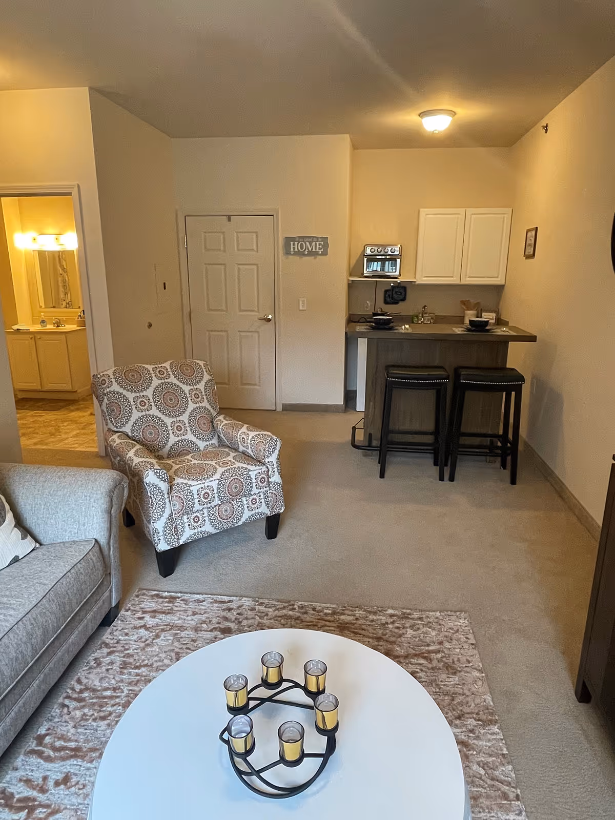 Open-plan living area with a sofa and patterned armchair around a coffee table, and a small kitchenette with bar stools in the background.