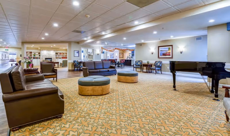 Spacious senior living common lounge with leather sofas, round ottomans, a grand piano, and patterned carpet.