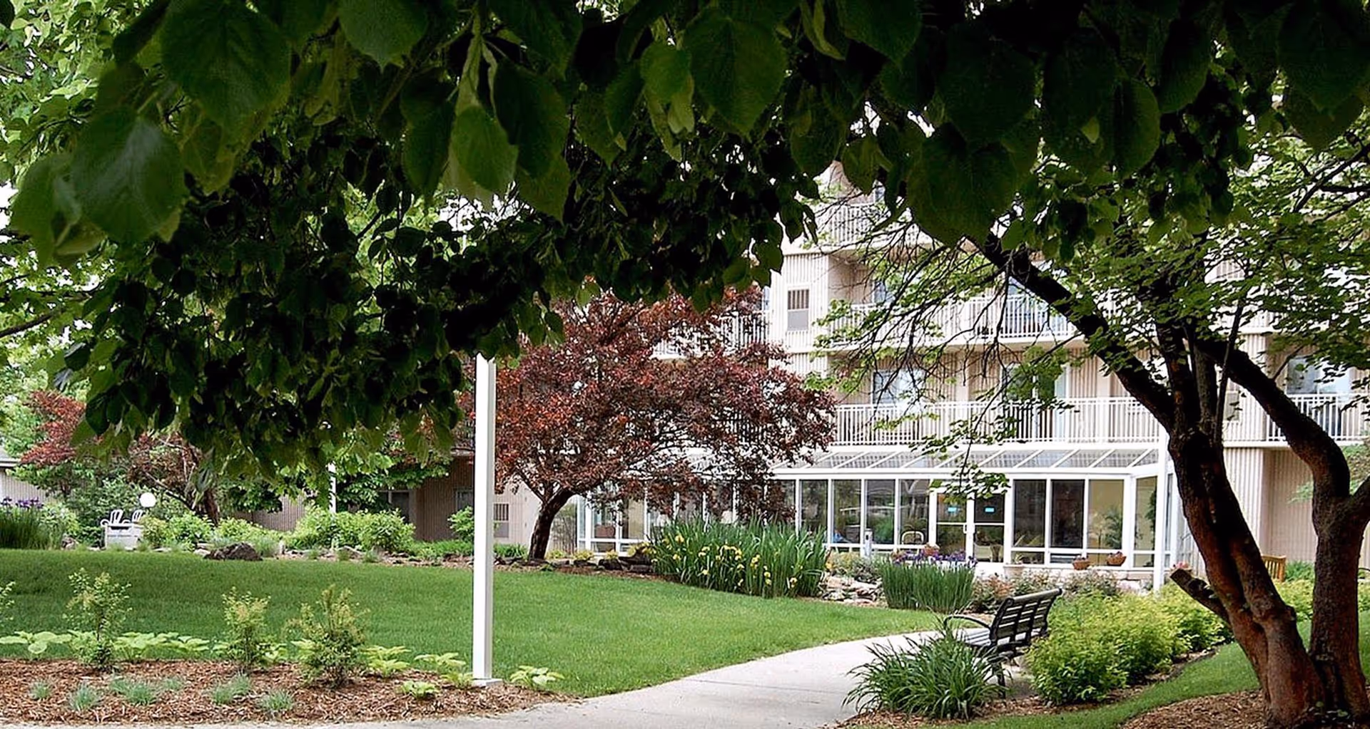 A landscaped outdoor area with a curved concrete pathway, green grass, various bushes, and trees. There are benches along the path and a multi-story building with balconies and large windows in the background.