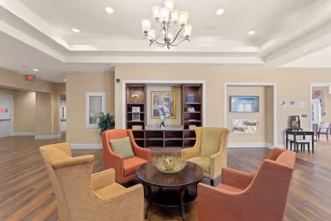 A bright and spacious common area in a senior living facility featuring four upholstered armchairs arranged around a round wooden coffee table with a decorative centerpiece. The room has wood flooring, beige walls, a built-in shelving unit with books and artwork, and a chandelier hanging from a tray ceiling. There are doorways leading to other rooms and a small table with a coffee machine in the background.