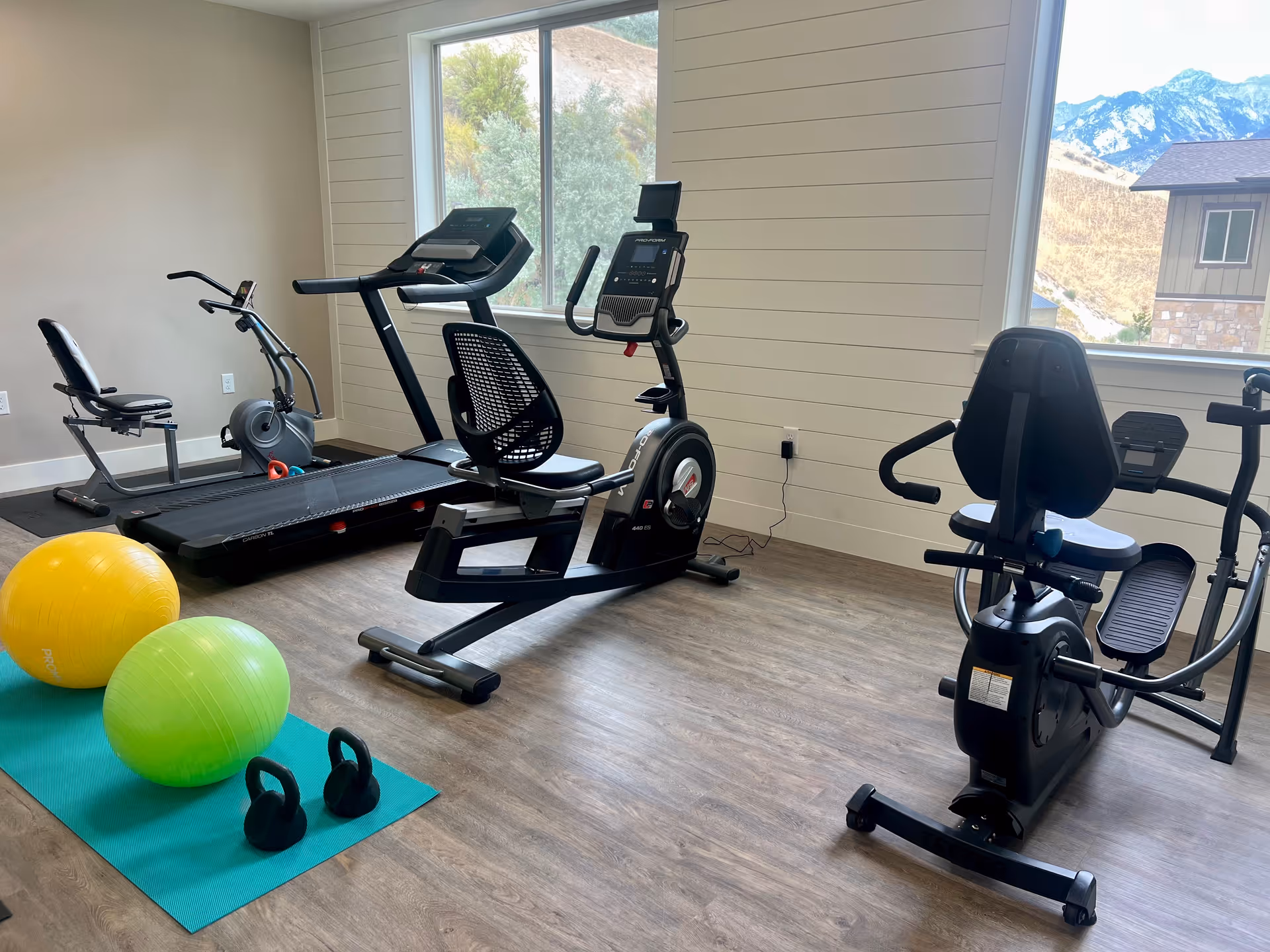 A senior living facility exercise room with various fitness equipment including a treadmill, two recumbent stationary bikes, two kettlebells, and two exercise balls on a blue mat. The room has large windows showing a view of mountains and a neighboring building.