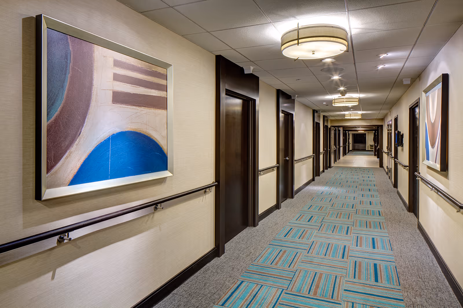 Well-lit interior hallway with patterned carpet, handrails, doors, and framed abstract artwork.