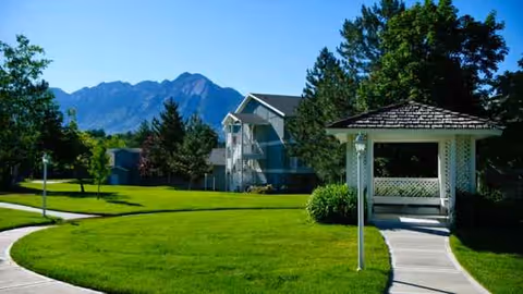 Green lawn, winding sidewalk, gazebo, and blue multi-unit building with mountains in the background.
