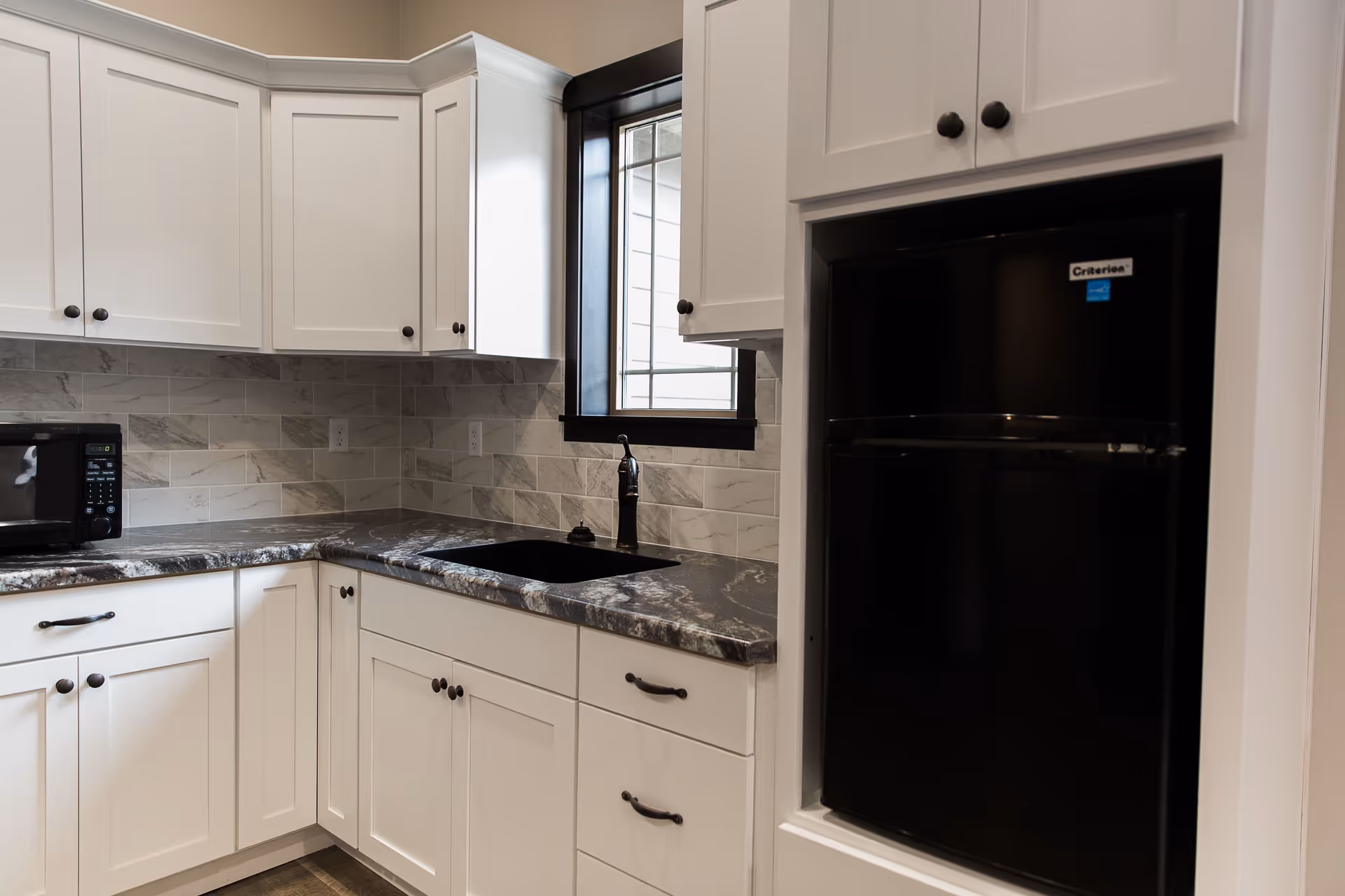 A modern kitchen corner with white cabinets, a black countertop, a black sink with a faucet, a black microwave on the counter, and a black refrigerator built into the cabinetry. There is a window with a black frame above the sink and a tiled backsplash in shades of gray and white.