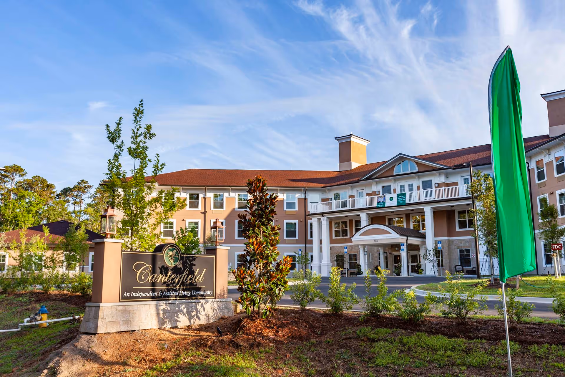Exterior view of Canterfield of Tallahassee, a multi-story senior living facility with a beige and white facade, a red roof, and a covered entrance. The foreground features a landscaped area with a sign displaying the facility's name and description, along with a tall green flag on the right side. The sky is clear with some wispy clouds.