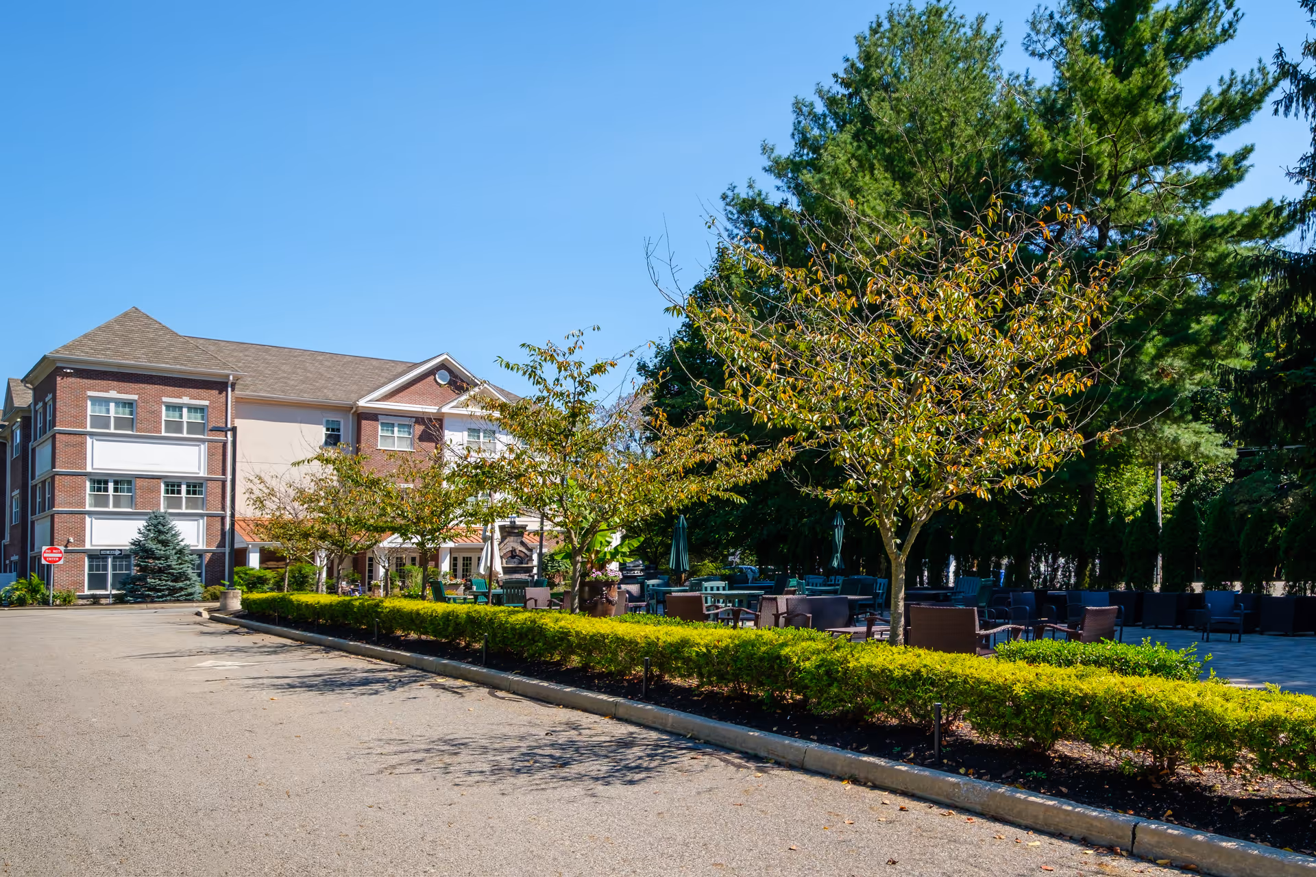 Outdoor patio area at Brandywine Mahwah by Monarch featuring multiple seating arrangements with chairs and tables surrounded by trees and greenery, adjacent to a three-story brick and beige building under a clear blue sky.