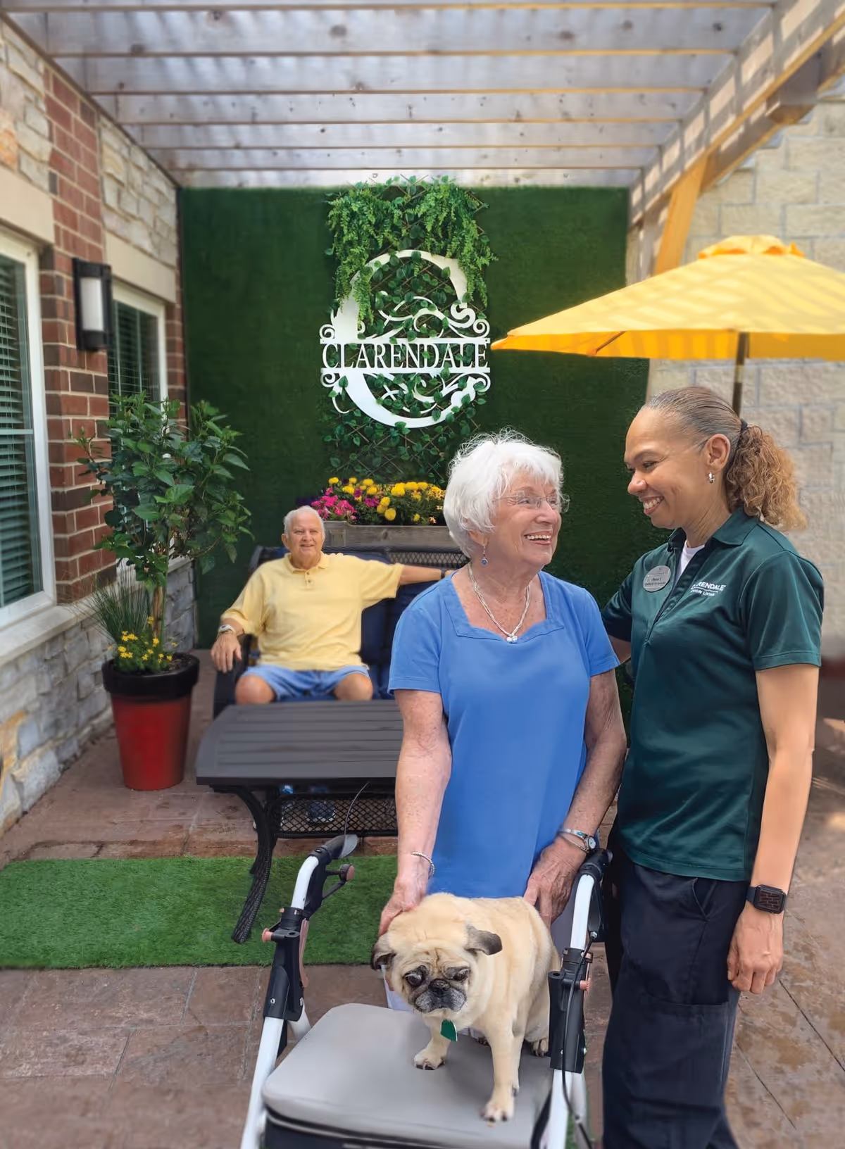An elderly woman with white hair and glasses stands with a walker that has a small pug dog sitting on it. She is smiling and looking at a female staff member wearing a green Clarendale uniform shirt. In the background, an elderly man in a yellow shirt and blue shorts sits on a bench under a pergola with a green wall and a Clarendale sign adorned with plants and flowers. A yellow umbrella is also visible.