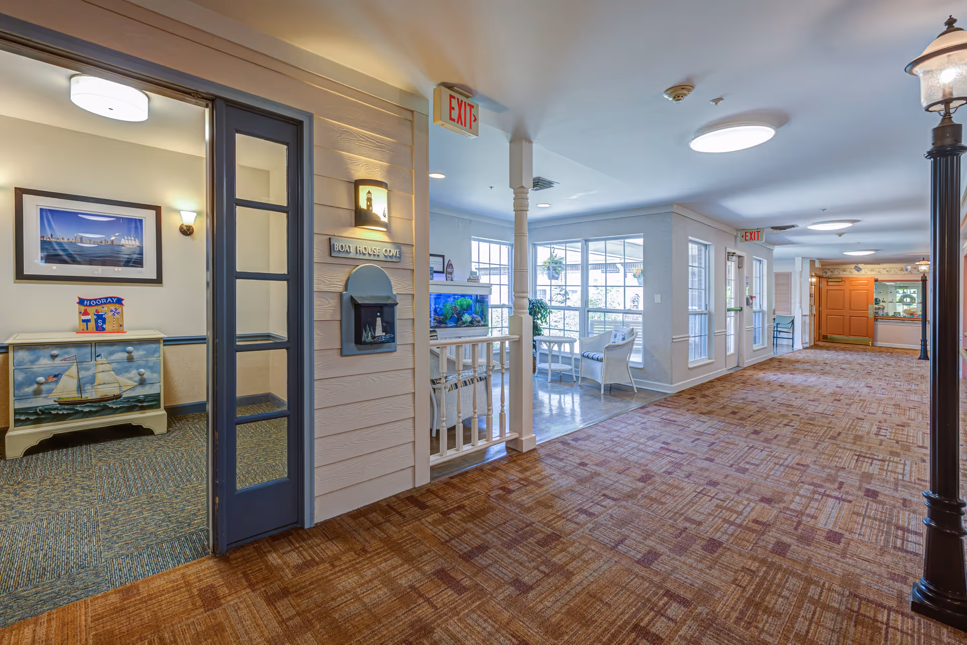 A brightly lit hallway in a senior living facility with patterned carpet and white walls. To the left, there is a room with a nautical-themed dresser and a framed picture on the wall. The hallway features a small seating area with white wicker chairs and a table near large windows. There are exit signs visible on the ceiling, and a decorative lamp post stands on the right side of the hallway.