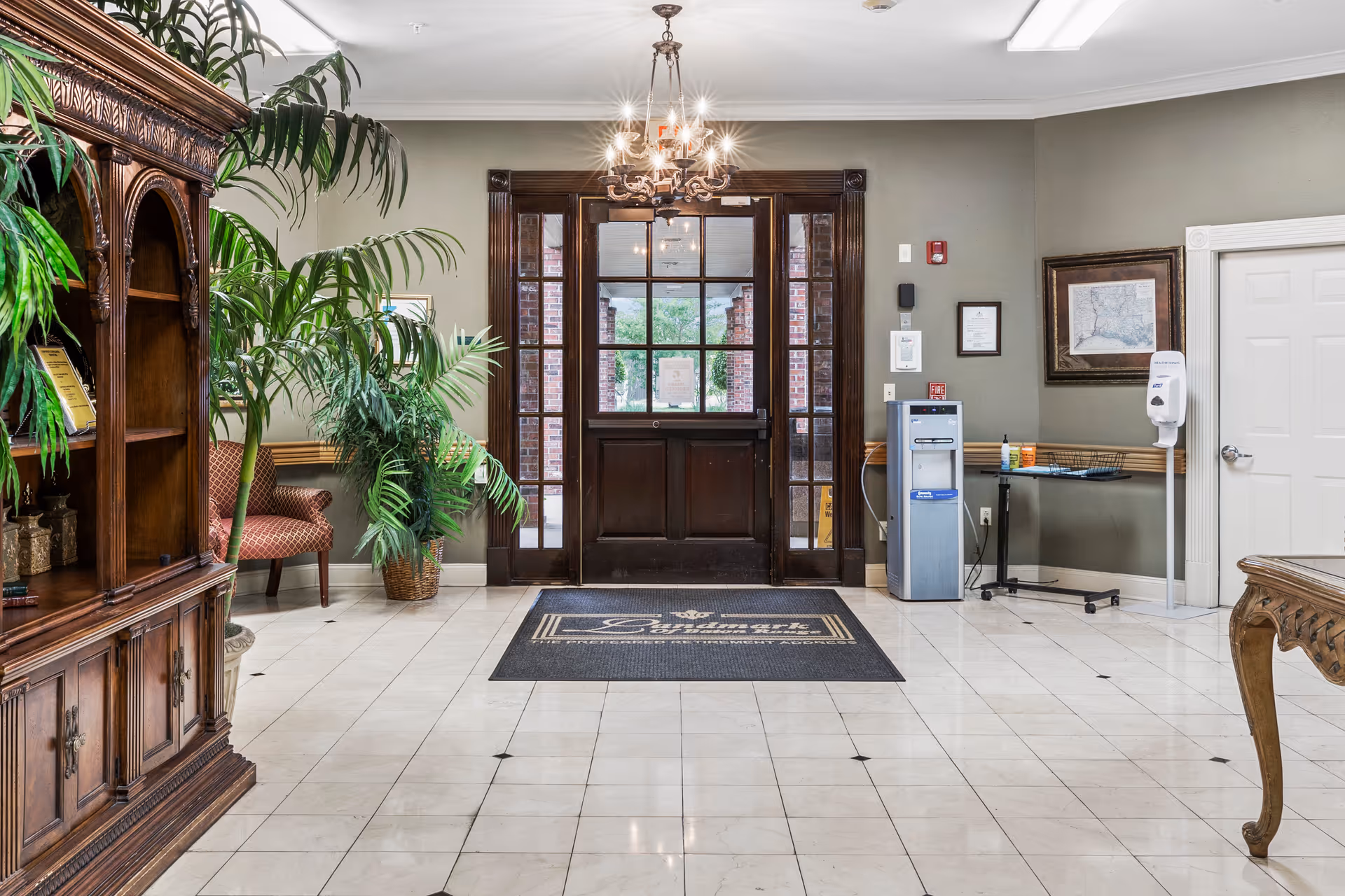 Entrance lobby of a senior living facility with a dark wooden door featuring glass panels, a chandelier hanging from the ceiling, a large indoor plant, a wooden cabinet on the left, a water dispenser, and a hand sanitizer station on the right. The floor is tiled with light-colored tiles and a dark mat with the word 'Landmark' is placed in front of the door.