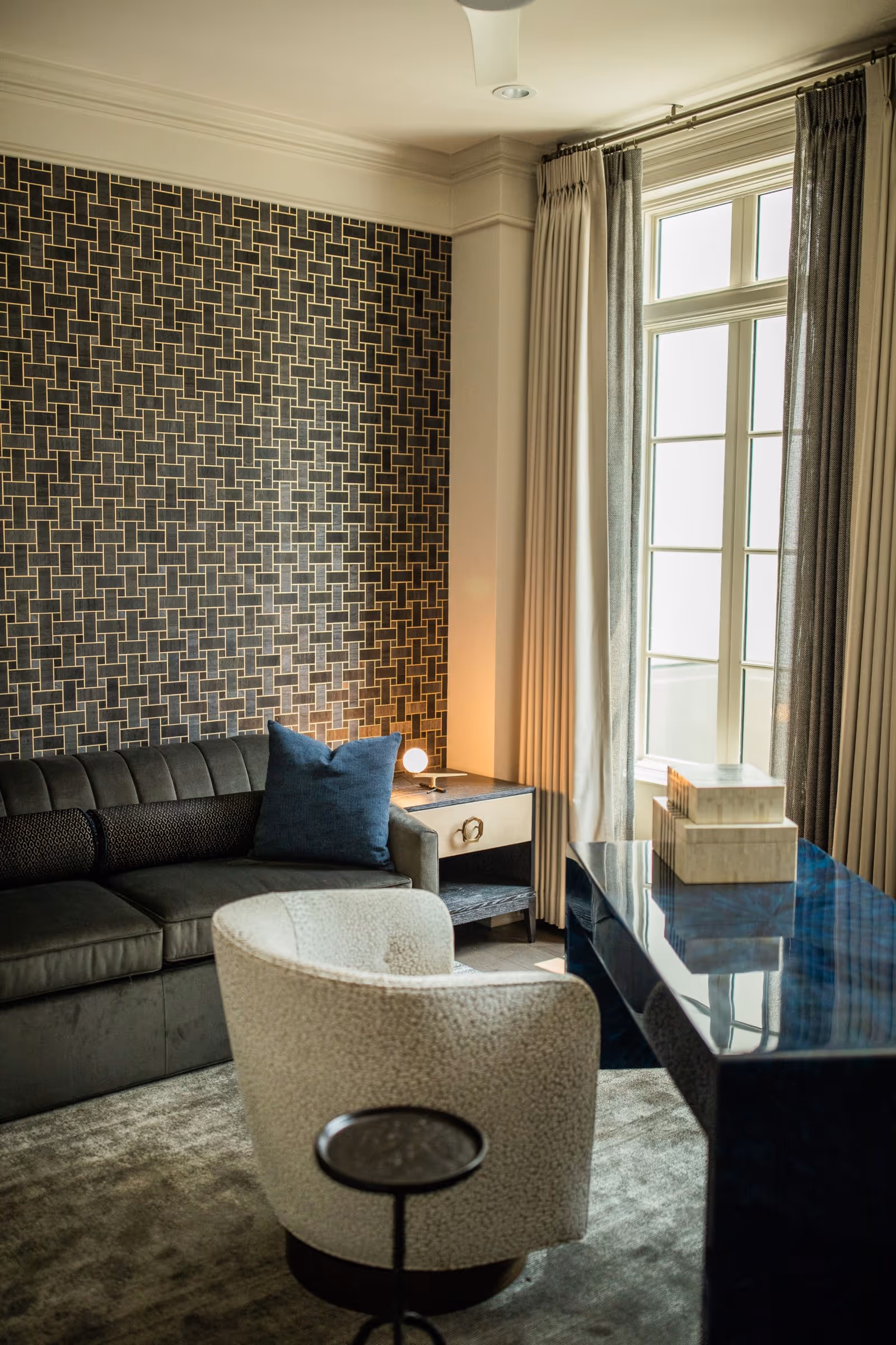 A cozy interior room featuring a dark gray sofa with a blue pillow, a small side table with a round lamp, a white textured armchair, a glossy dark blue desk with stacked decorative boxes, and a large window with beige curtains allowing natural light to fill the space. The wall behind the sofa has a geometric patterned wallpaper.