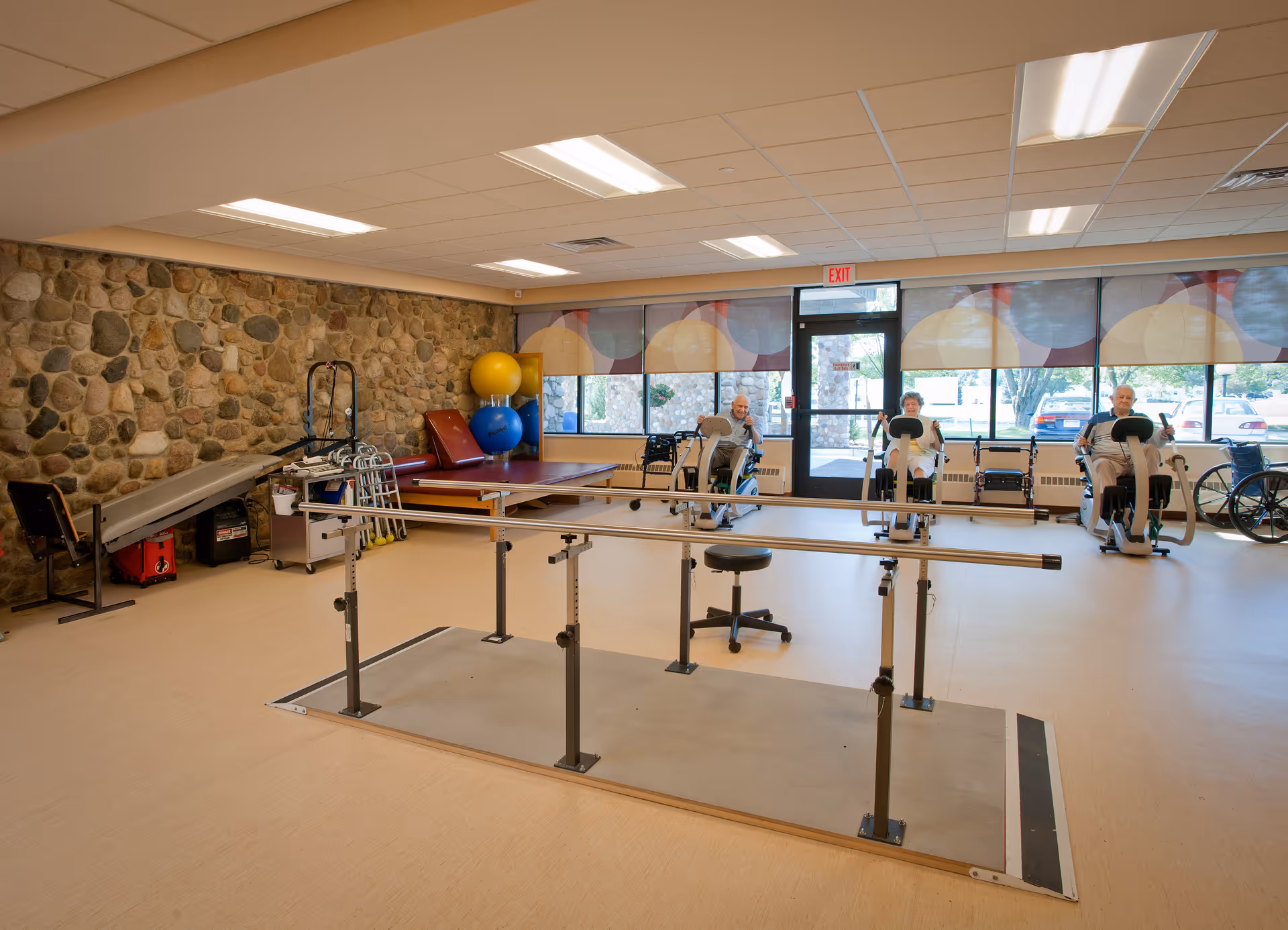 A spacious rehabilitation or physical therapy room in a senior care center with parallel bars for walking exercises in the center. Three elderly individuals are using stationary exercise bikes near large windows with patterned blinds. The room has a stone accent wall, exercise mats, therapy balls, walkers, and a wheelchair visible.