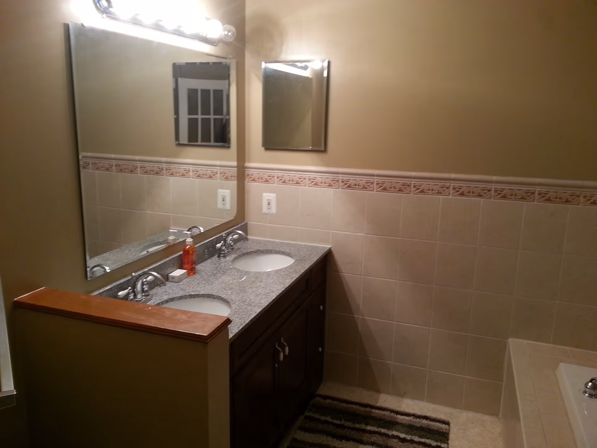 Bathroom interior with a double sink vanity featuring a granite countertop, two mirrors above the sinks, beige tiled walls with a decorative border, a striped rug on the floor, and a bathtub partially visible on the right side.