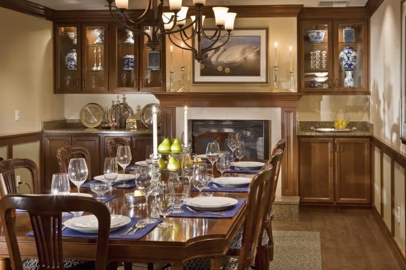 A formal dining room with a long wooden table set for a meal, including plates, glasses, silverware, and blue placemats. The room features wooden cabinetry with glass doors displaying decorative items, a fireplace with a framed picture above it, and a chandelier overhead providing warm lighting.