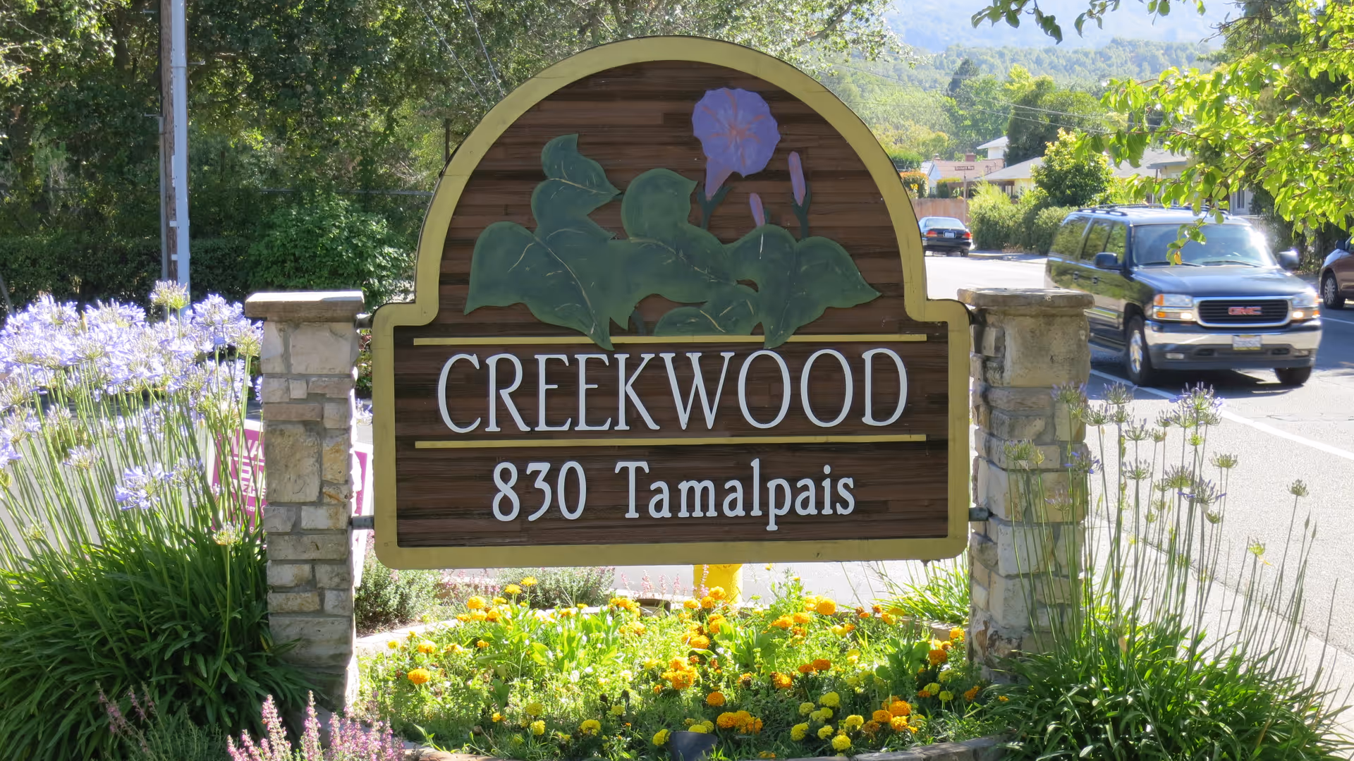 A wooden sign for Creekwood Care Facility at 830 Tamalpais, surrounded by flowers and greenery, with a street and cars in the background.
