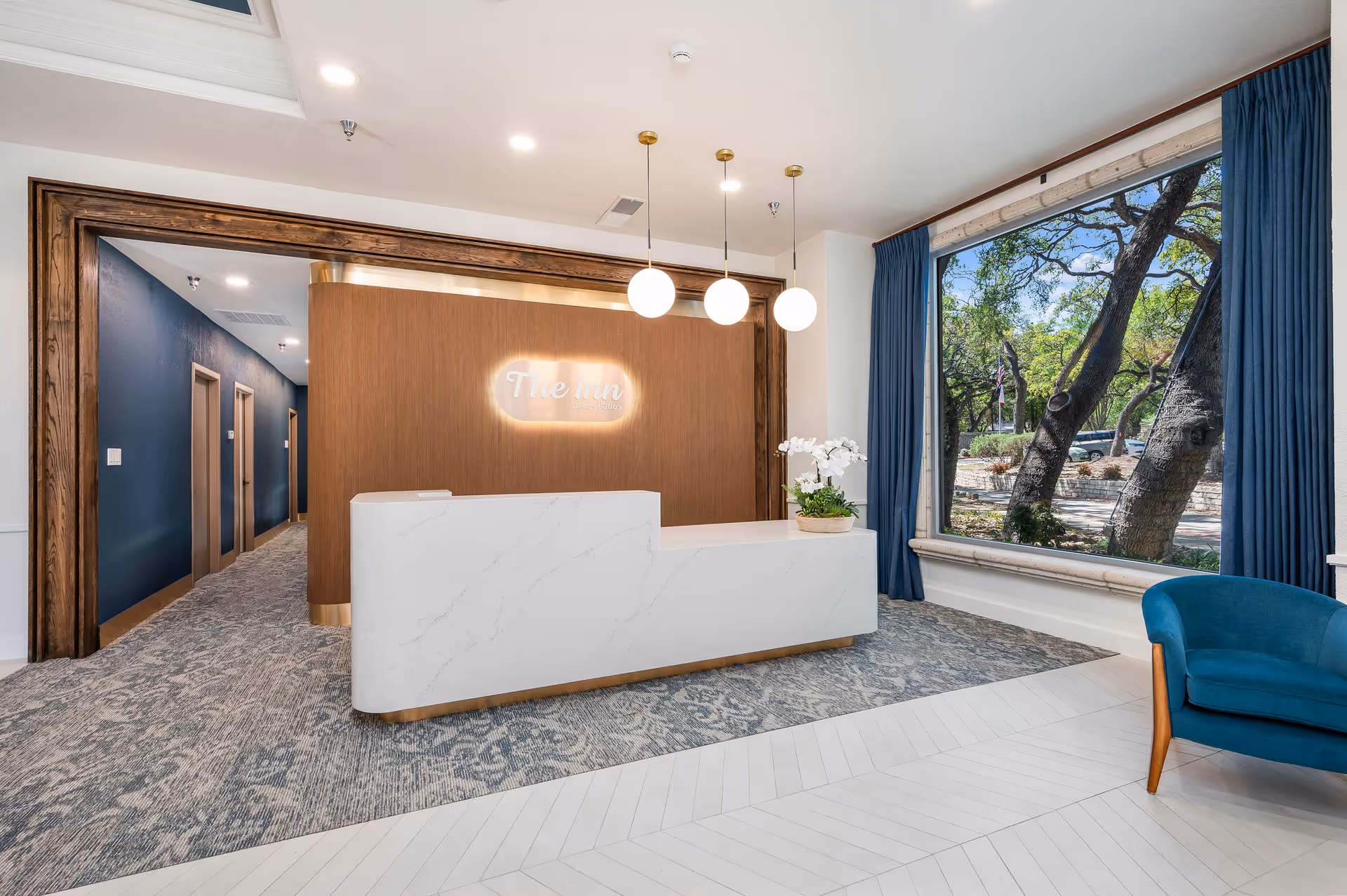 Reception area of The Inn at Los Patios featuring a modern white marble front desk with a flower arrangement, three hanging globe pendant lights, a large window with blue curtains showing trees outside, and a blue armchair on the right side.