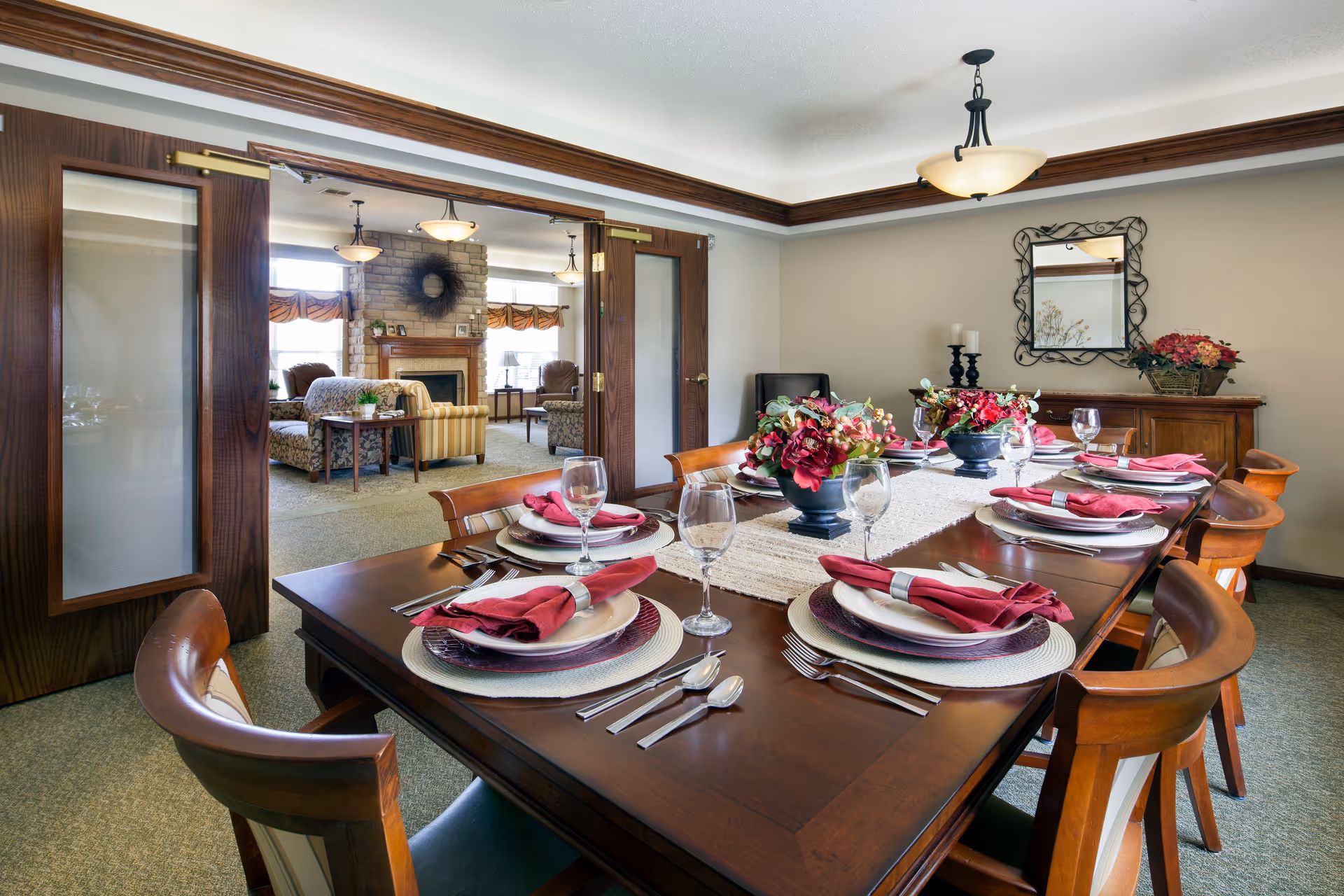 A dining room set for a meal with a large wooden table, eight chairs, and place settings including plates, silverware, wine glasses, and red cloth napkins. The table has two floral centerpieces. In the background, there is a living room area with sofas, armchairs, a stone fireplace, and large windows letting in natural light. The dining room has a decorative mirror on the wall and a light fixture hanging from the ceiling.