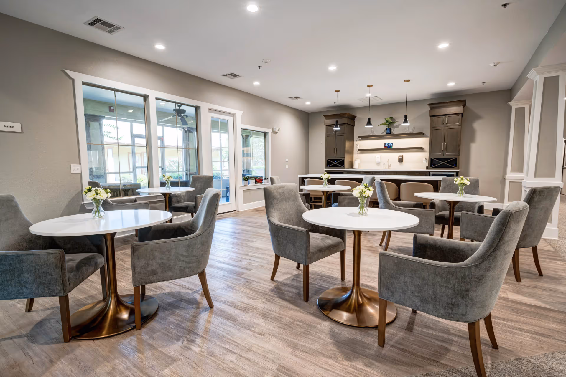 A modern dining area with round white tables and gray upholstered chairs. Each table has a small vase with white flowers. The room features wood flooring, large windows, and a counter with cabinets and pendant lights in the background.