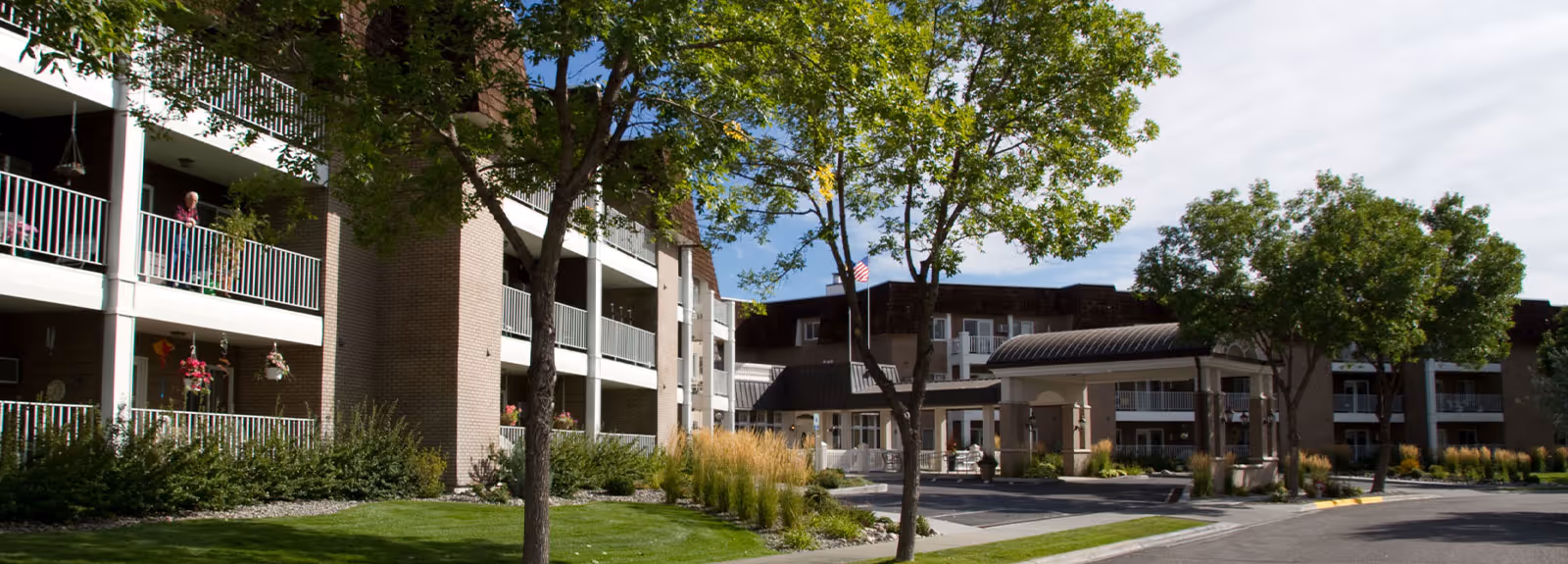 Exterior view of Westpark Village senior living facility showing a multi-story building with balconies, trees, and landscaped greenery under a partly cloudy sky.
